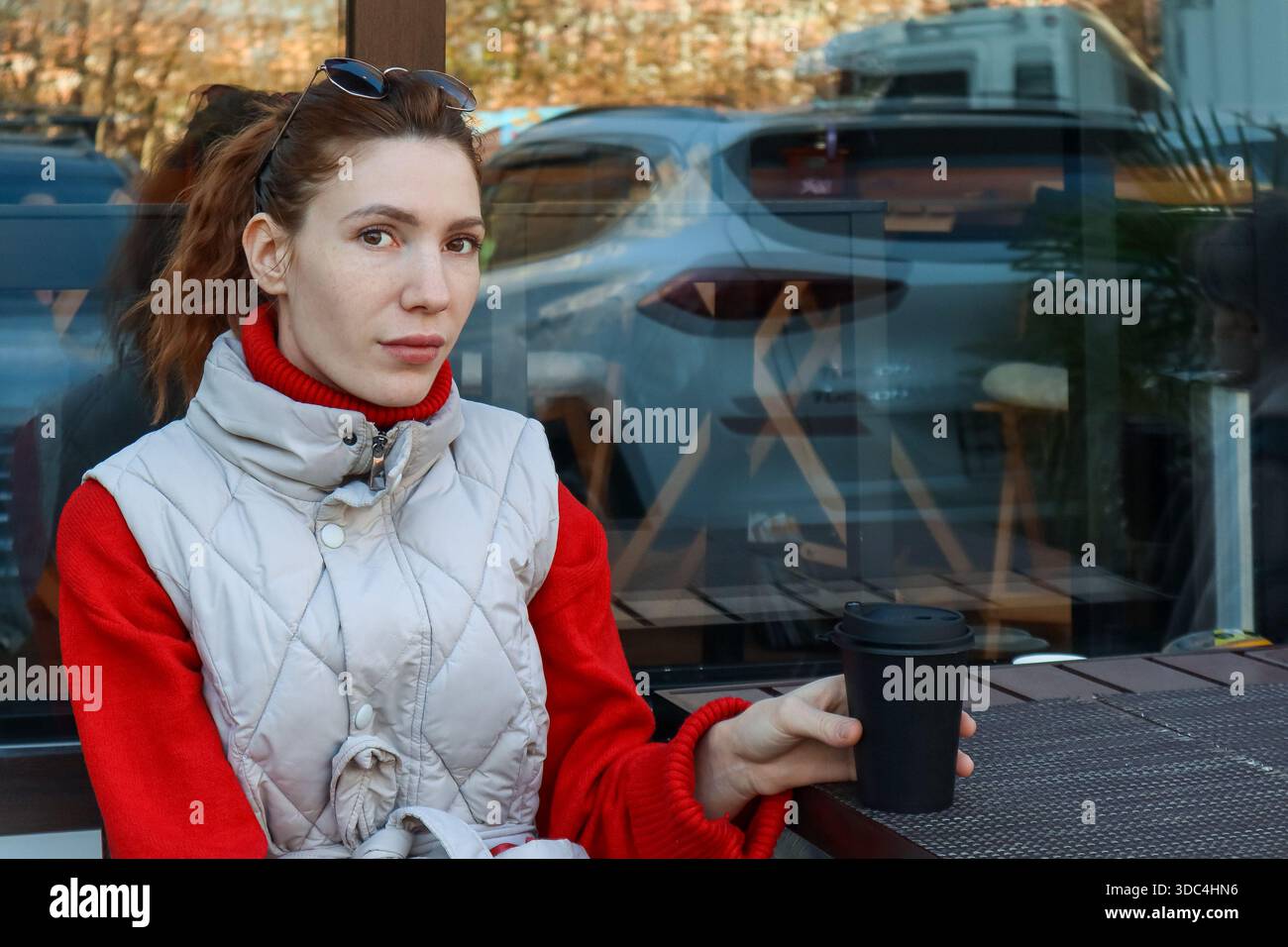 Una giovane donna in maglione rosso e gilet grigio siede al tavolo del caffè tenendo una tazza nera da asporto, guardando con attenzione. Foto Stock