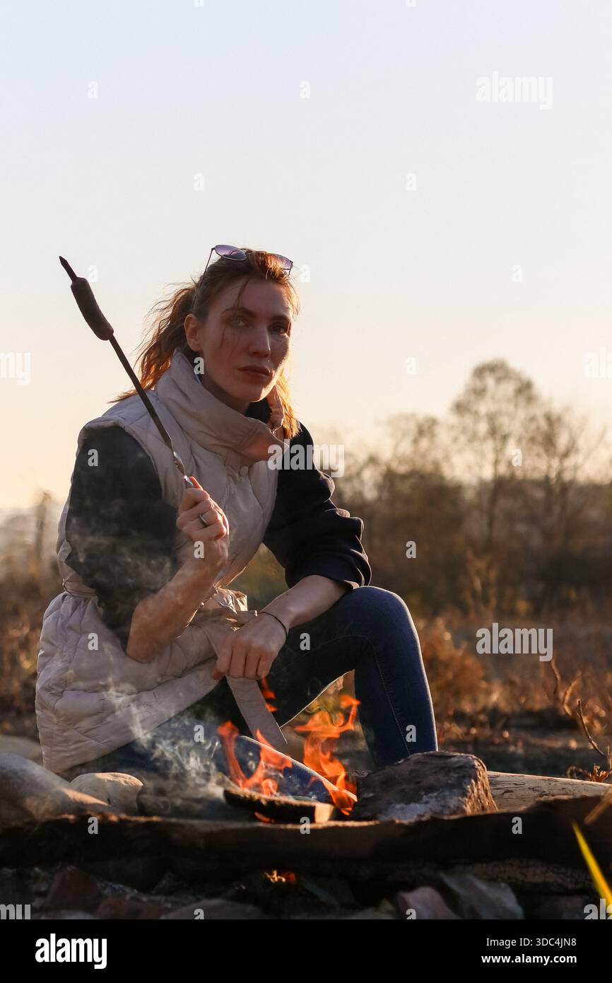Donna in giubbotto leggero estende un bastone con marshmallow nelle fiamme del fuoco, seduto a terra in un campo autunnale con alberi lontani e cielo blu (102) Foto Stock