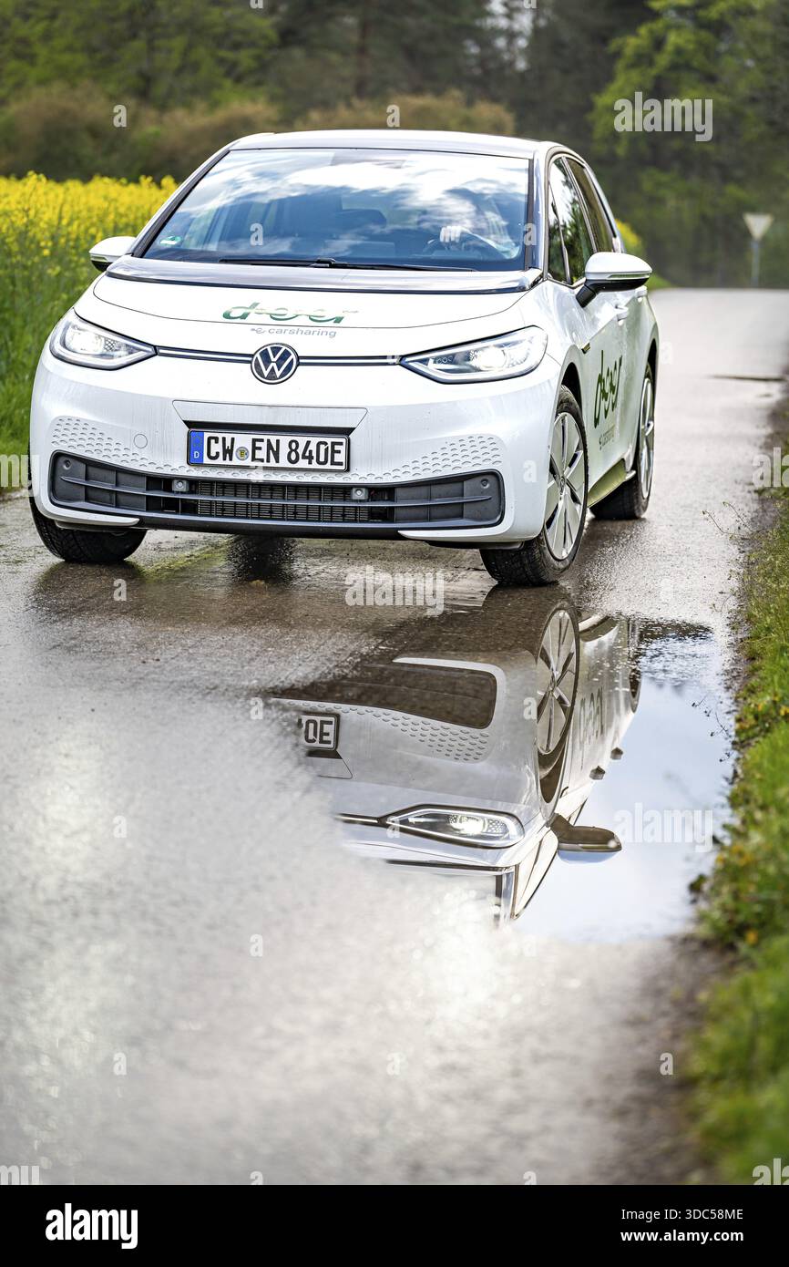 Auto su strada bagnata con un chiaro riflesso nella pozzanghera, immersa nel verde, ID3 VW, condivisione auto con cervi. Calw, Germania Foto Stock