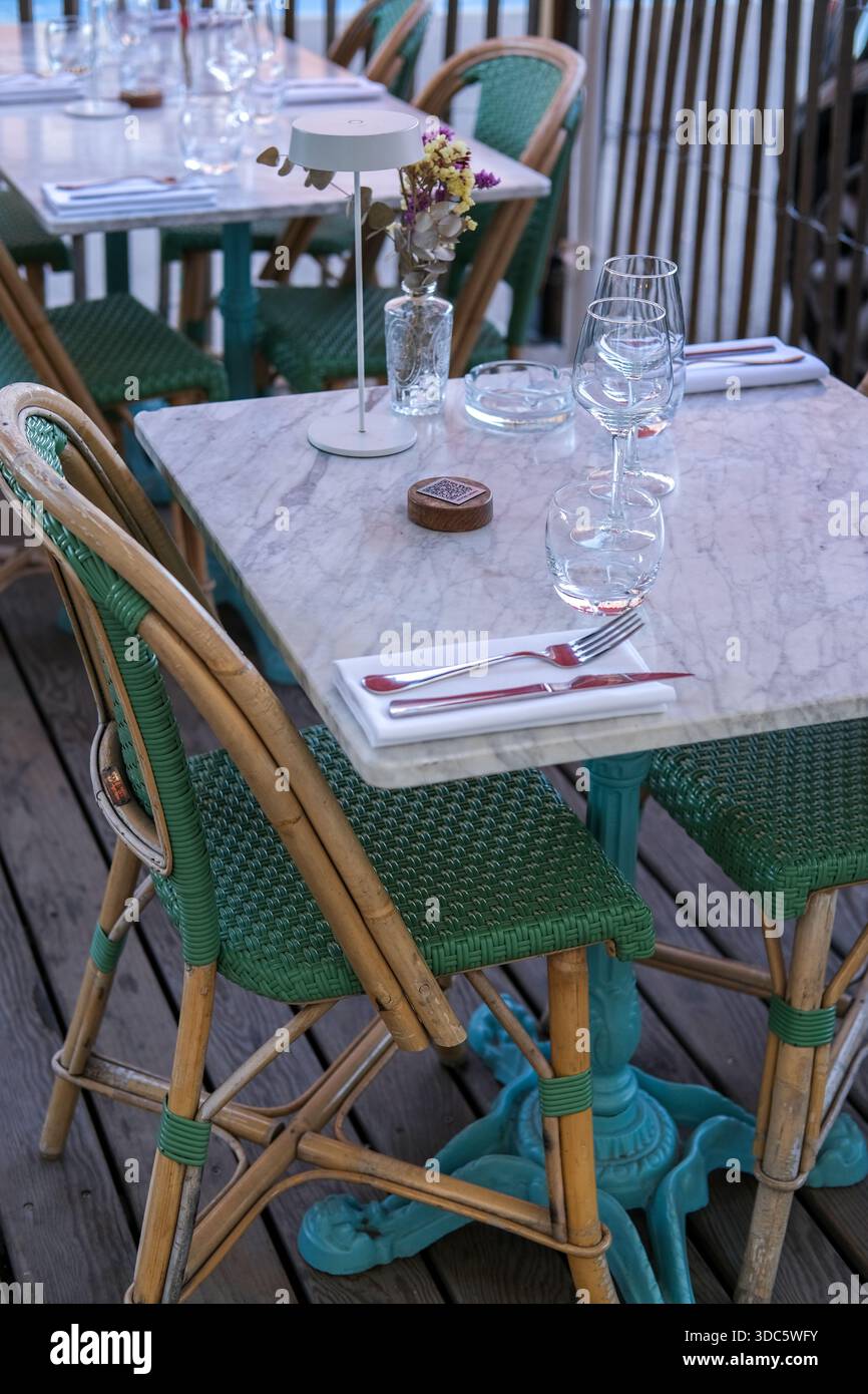 Tavolino da caffè con piano in marmo, bicchieri da vino, bicchieri da acqua, posate, tovagliolo, piccola lampada e vaso, sedie in vimini, terrazza in legno. Foto Stock