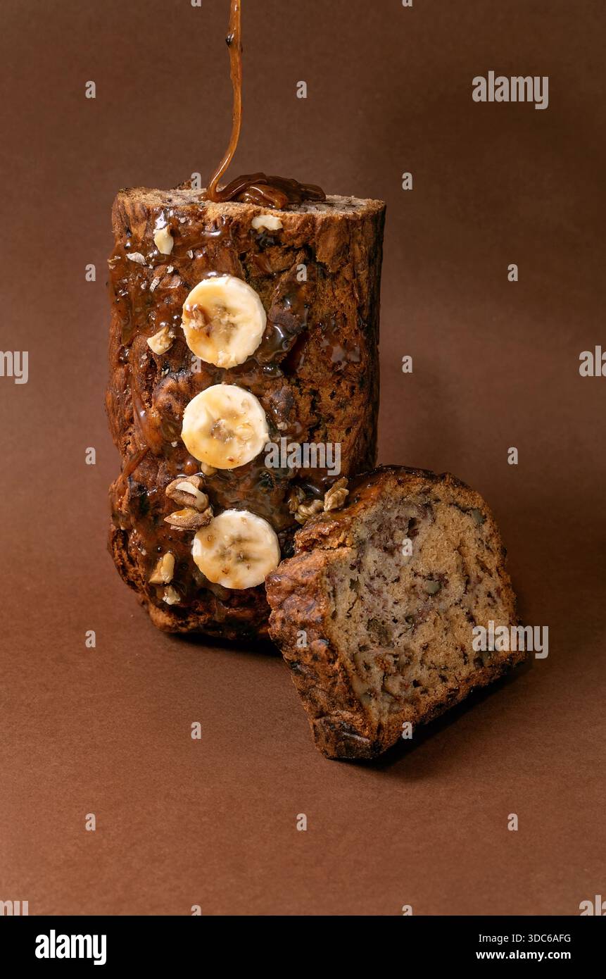 Natura morta moderna con pane alla banana fatto in casa. Pagnotto con noci, condito con caramello, in posizione verticale su sfondo marrone. Salsa versata dall'alto Foto Stock
