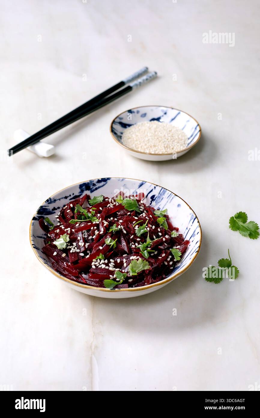 Insalata di barbabietole in stile coreano. Barbabietola grattugiata marinata con semi di sesamo e foglie di coriandolo in un recipiente di ceramica. Recipiente vicino con semi di sesamo e bacchetta Foto Stock