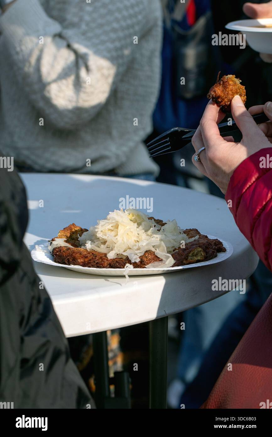 Persona che mangia il tradizionale fast food ungherese ceco fritto, pane piatto bramboraky con impasto di patate fritto con crauti su piatto di carta anche all'aperto Foto Stock