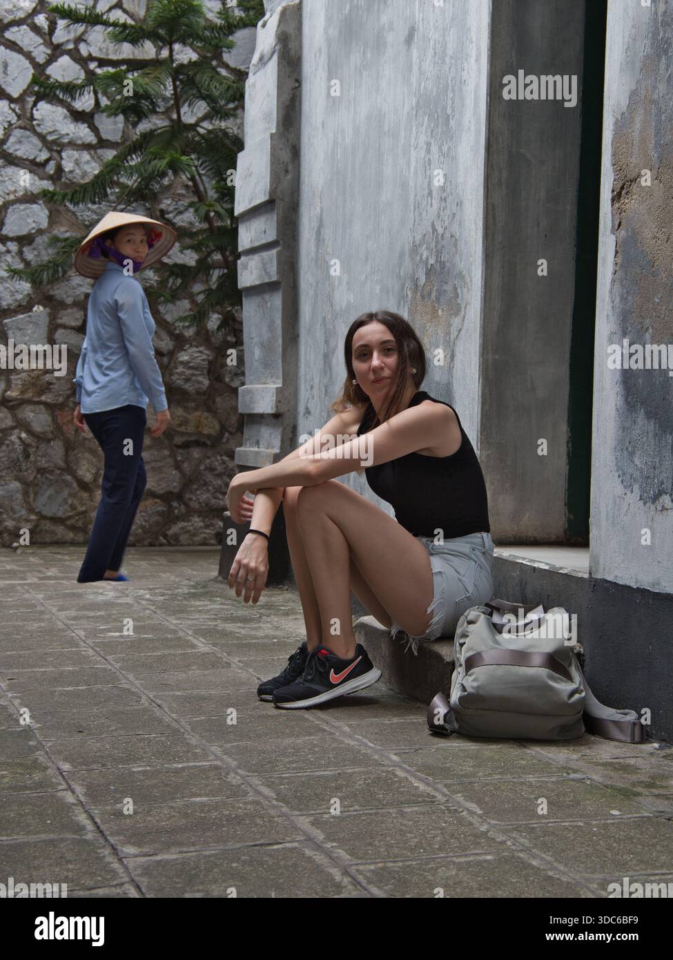hanoi-Vietnam-9 agosto 2019, Un turista riposa su gradini vicino a un muro intemprato. Una donna vietnamita locale che indossa un tradizionale cappello conico (nón lá) cammina Foto Stock