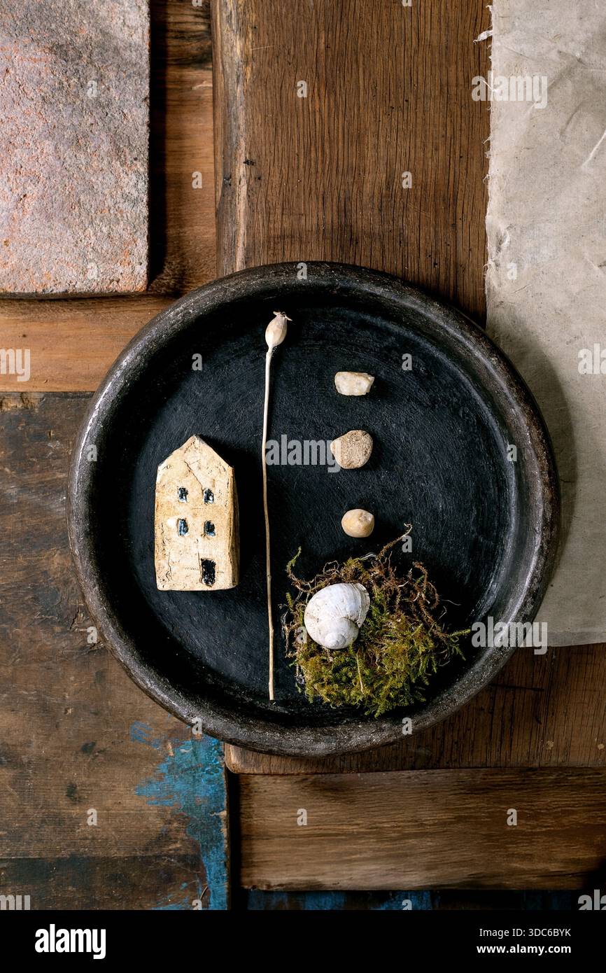 Primo piano recipiente in ceramica nera con una casa in ceramica in miniatura, piccoli ciottoli, nido di muschio con guscio su fondo rustico in legno. Materiali naturali artistici Foto Stock