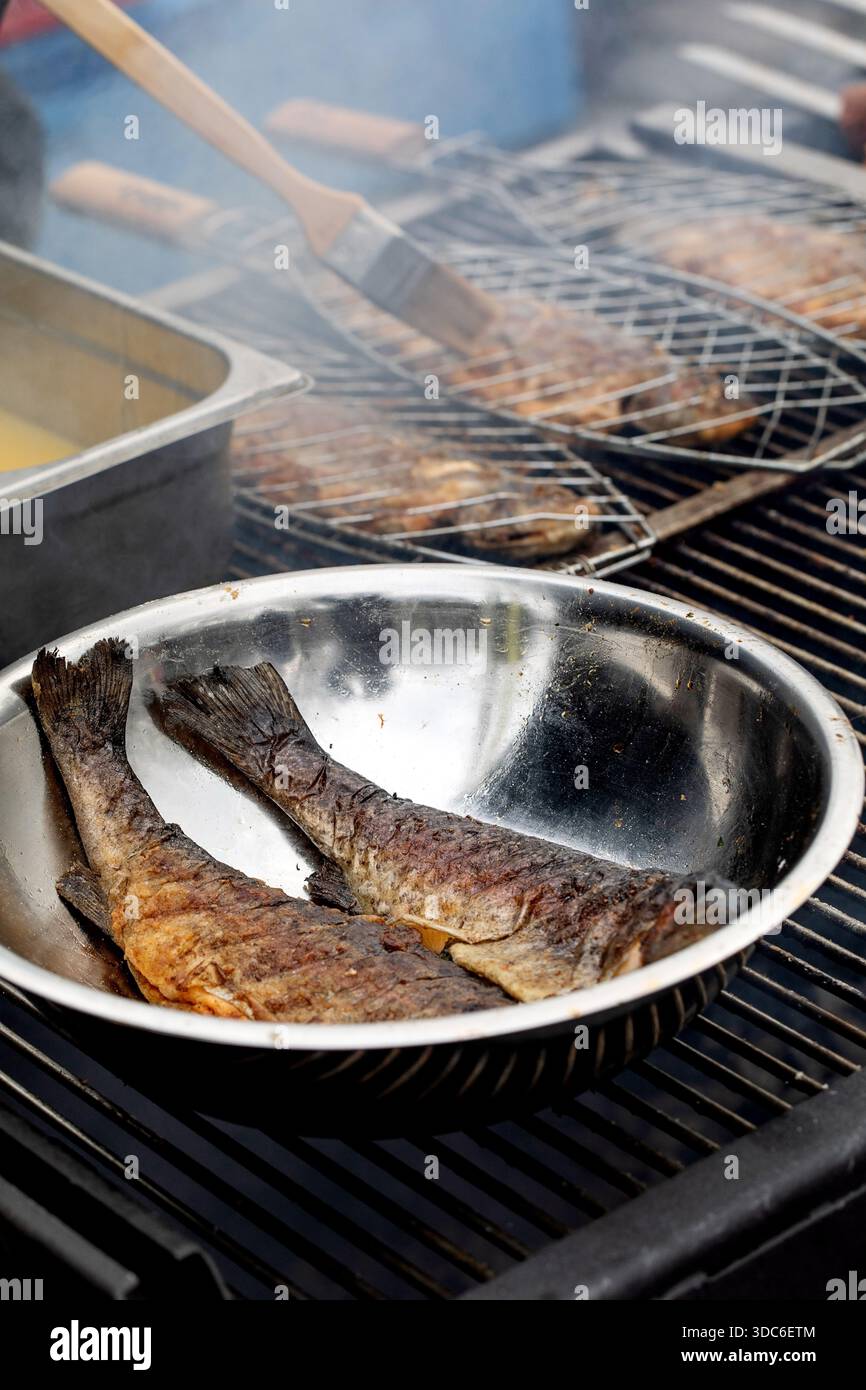 Code di pesce fresco grigliate al barbecue. Recipiente in metallo con parti di pesce cotte sulla griglia. Processo di cottura con pennello che applica la salsa sullo sfondo. Foto Stock