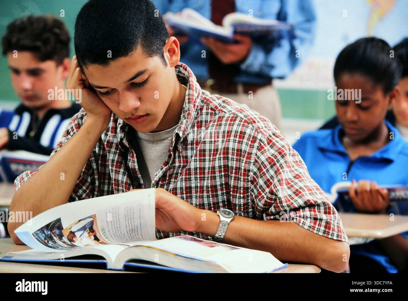 Diversi studenti della scuola si siedono alle scrivanie in una classe. Un Latinostudent si concentra su un libro mentre altri leggono. L'atmosfera mostra l'apprendimento e lo studio. Foto Stock