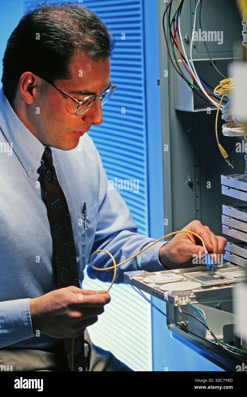 Un tecnico si concentra sulla creazione di connessioni all'interno di un server rack. Sta indossando occhiali e una cravatta. I fili sono visibili quando gestisce i componenti del computer Foto Stock