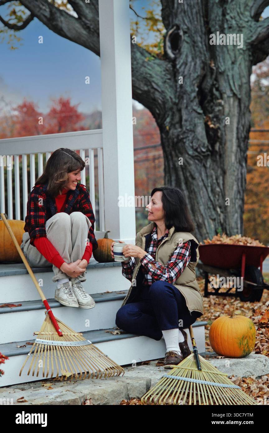 Due donne amiche si siedono sui gradini di una casa, impegnandosi in una conversazione. Sono circondati da zucche e rastrelli, il che indica che è autunno nel gar Foto Stock
