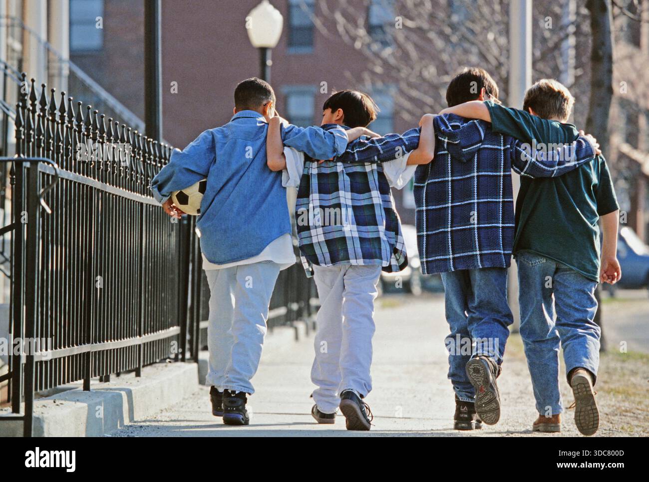 Quattro ragazzi camminano fianco a fianco su un marciapiede in città. Indossano abiti casual e uno di loro tiene una palla da calcio. Sembra che a loro piaccia ogni othe Foto Stock