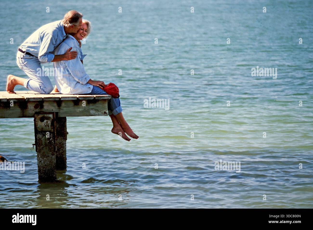 La coppia senior siede su un molo di legno sopra l'acqua. Sorridono e condividono un momento mentre i loro piedi sfrecciano sopra la superficie. C'è il sole e sembrano andati Foto Stock