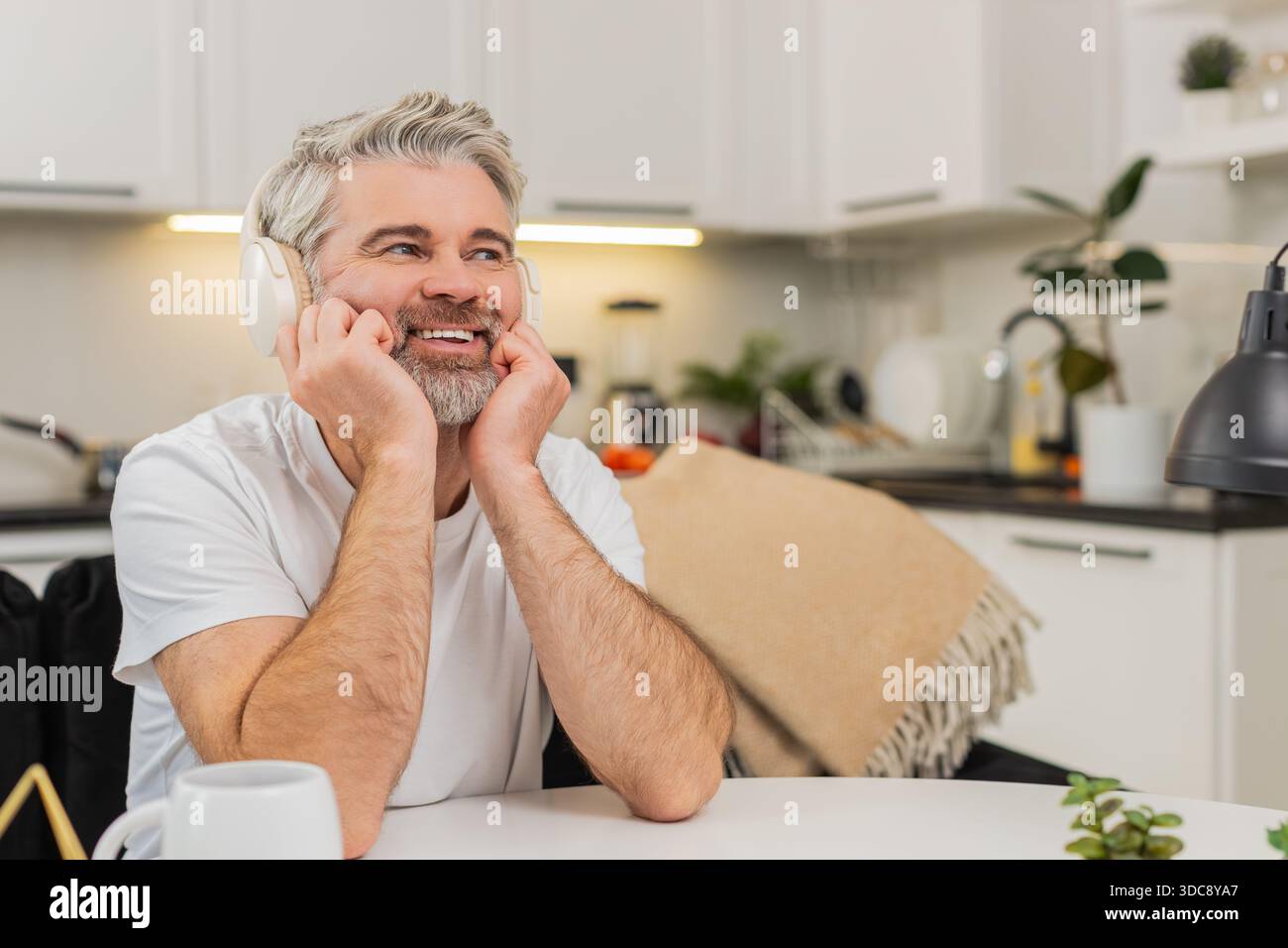 L'uomo di mezza età a casa ascolta la musica con le cuffie wireless si appoggia alle mani sorrisi e sogni. Un ragazzo maturo al tavolo sul divano pensa a una scelta responsabile il piano futuro si sente calmo e sicuro di sé. Foto Stock