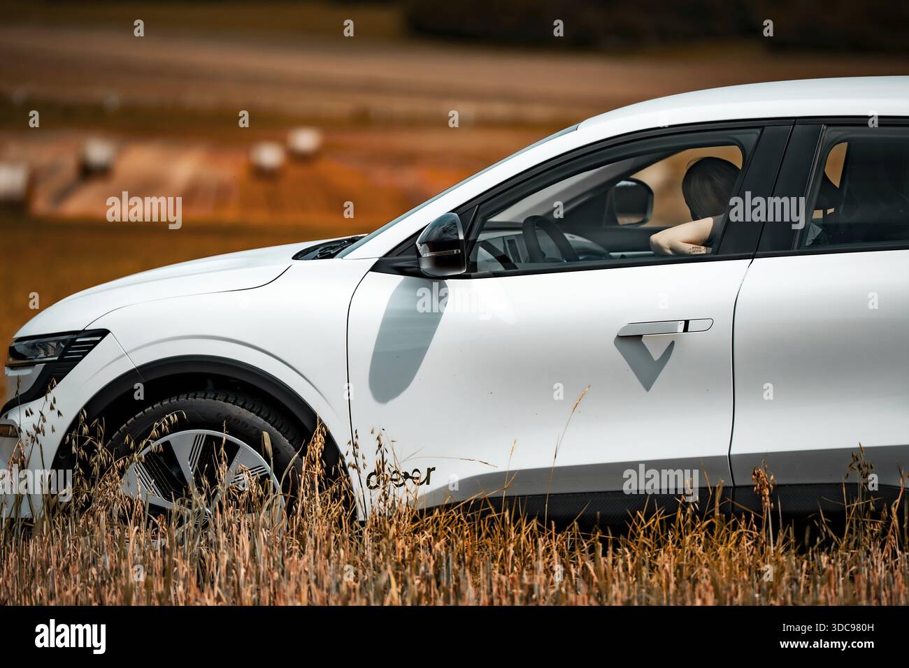 Auto elettrica bianca che attraversa un campo, conducente visibile, atmosfera estiva, condivisione di e-car con cervi, Renault Megane, Calw, Germania Foto Stock