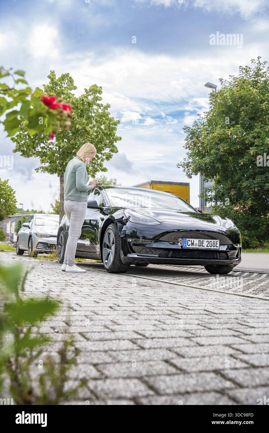 Persona in piedi accanto a un'auto nera su una strada asfaltata sotto il cielo blu con nuvole, Deer e-Carsharing, Tesla Model 3, Calw, Germania Foto Stock