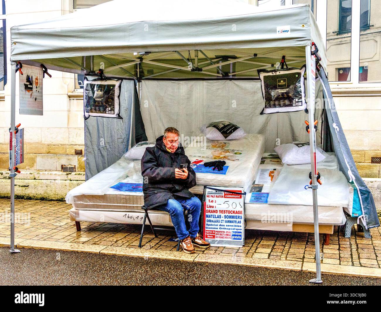 Il venditore di materassi che scorre al telefono nella "giornata lenta" al mercato all'aperto il giorno della pioggia - Loches, Indre-et-Loire (37), Francia. Foto Stock