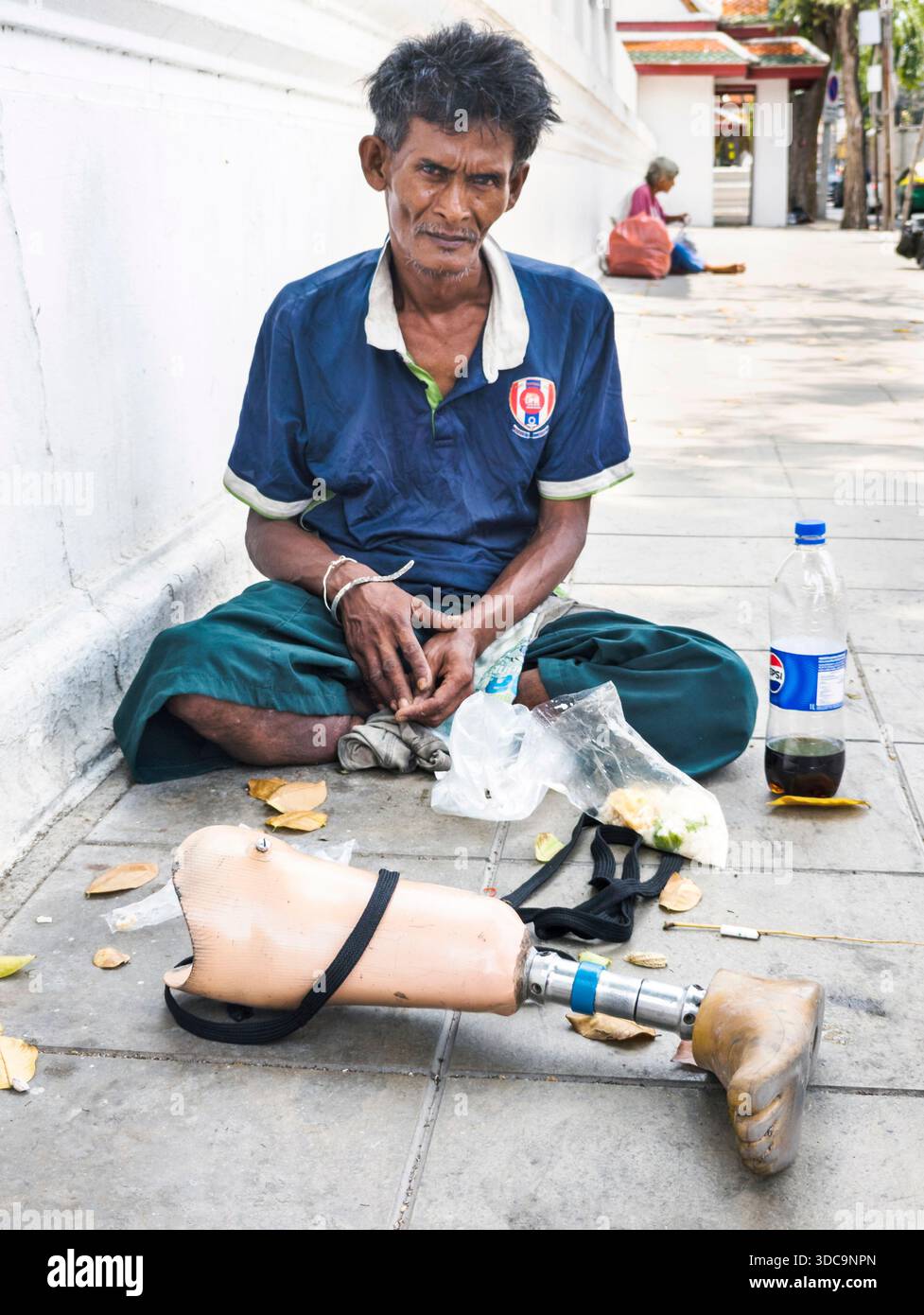 Bangkok, Thailandia - 21 aprile 2025: Un uomo senzatetto si siede sul marciapiede, la sua falsa gamba posta di fronte a lui, per riposarsi, su un caldo afterno Foto Stock
