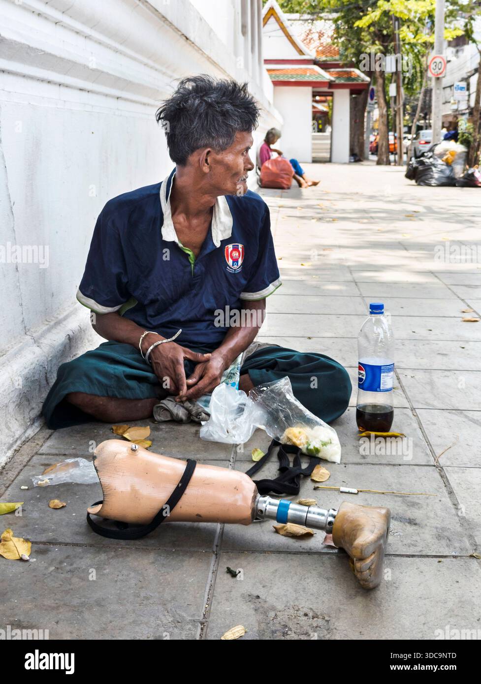 Bangkok, Thailandia - 21 aprile 2025: Un uomo senzatetto si siede sul marciapiede, la sua falsa gamba posta di fronte a lui, per riposarsi, su un caldo afterno Foto Stock