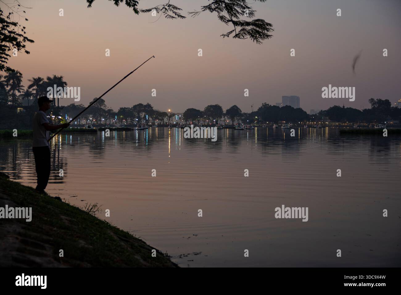 Silhouette Fisherman lancia una linea accanto a un lago calmo al tramonto con luci della città Foto Stock