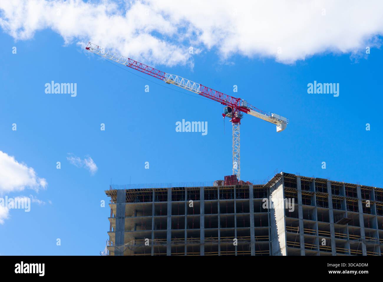 Alto edificio residenziale in costruzione con una grande gru a torre rossa e bianca in cima a un cielo blu luminoso con nuvole Foto Stock
