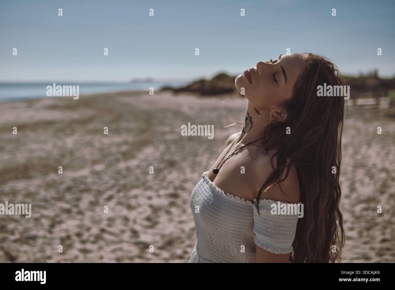 Giovane donna serena in abito bianco goditi il calore e il sole con gli occhi chiusi in piedi sulla spiaggia di sabbia, sentiti libero e felice. Momento meditativo Foto Stock