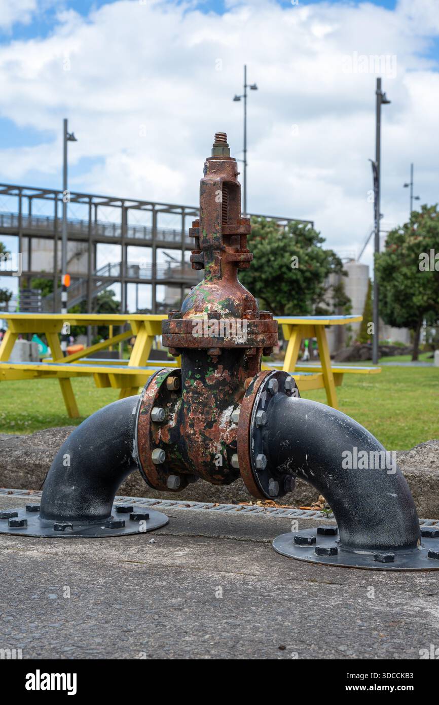 Una valvola dell'acqua in ferro d'epoca con vernice rossa sbucciante si trova su una conduttura arrugginita, mostrando anni di usura industriale Foto Stock
