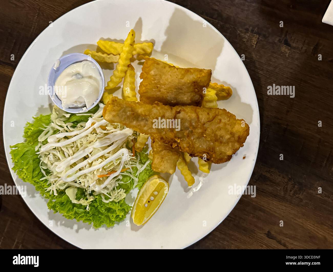 Filetti di pesce pastella dorati serviti su patatine croccanti tagliate a rughe con insalata di cavolo fresca e spicchi di limone su un piatto bianco in un ambiente accogliente. Foto Stock