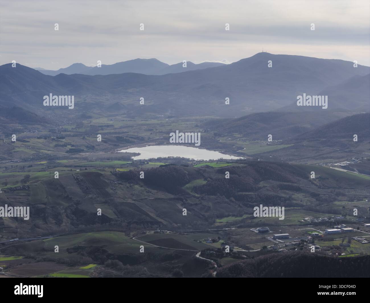 Vista aerea di un tranquillo lago incastonato tra colline ondulate e montagne lontane, un arazzo di verdi e blu tenui sotto una luce soffusa, po Foto Stock