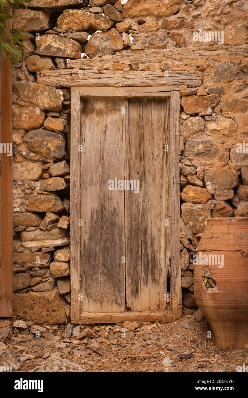 Porta di legno intemprata in rustico muro di pietra con vaso di argilla sull'isola fortezza di Spinalonga, Creta Grecia. Ingresso antico per la storica architettura veneziana. Foto Stock