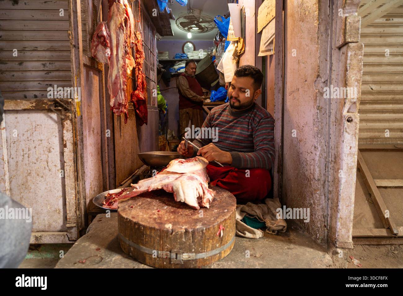 Lahore, Pakistan - 21 dicembre 2025: Veduta di un macellaio che prepara meticolosamente la carne, i tagli crudi sono aspri contro il blocco di legno usurato e il vivace Foto Stock