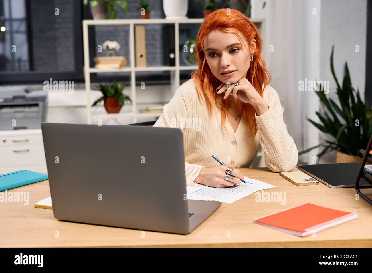 Una giovane donna dai capelli rossi brillanti si impegna con cura nelle sue attività presso una moderna scrivania da ufficio. Foto Stock