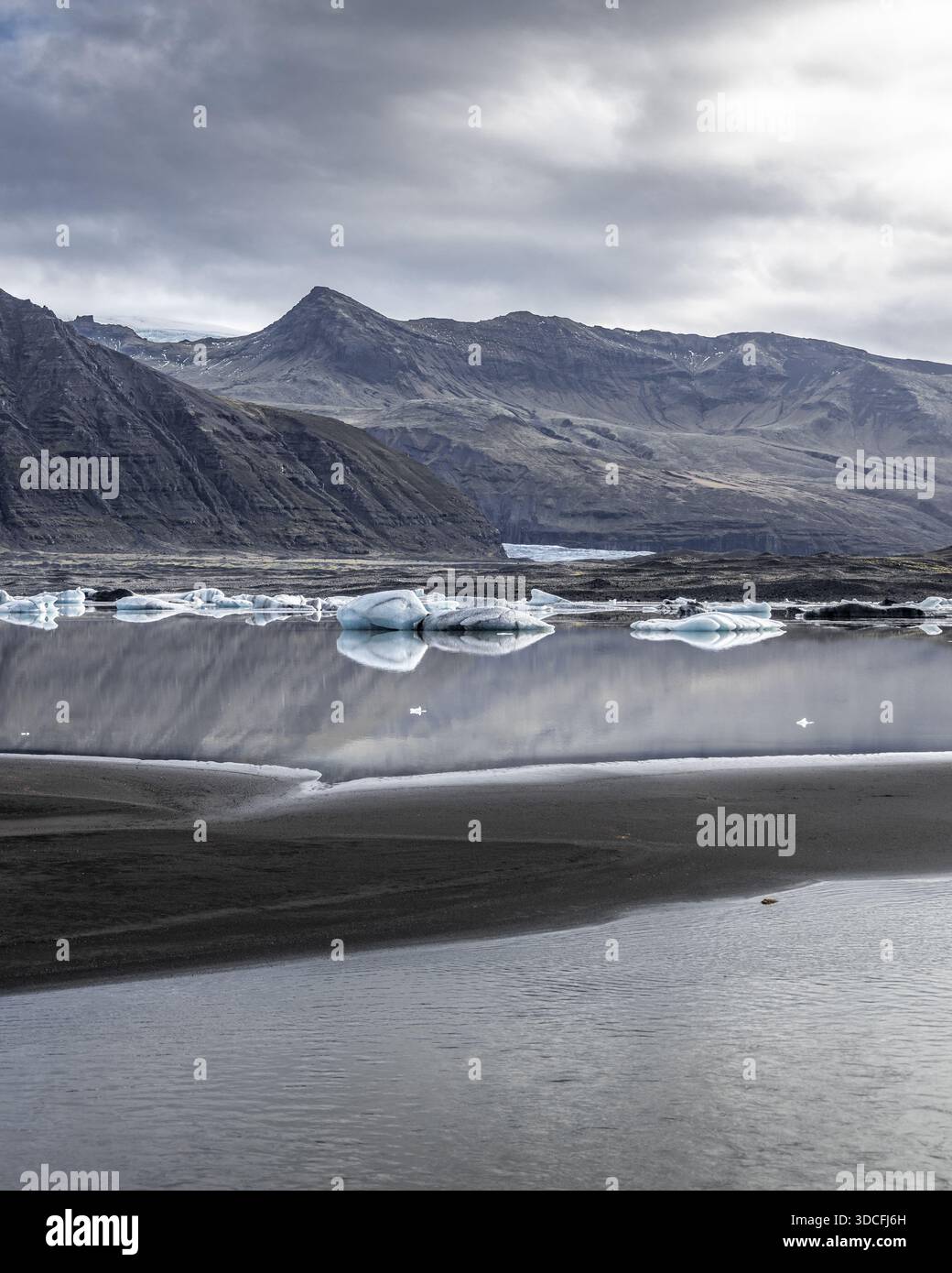 Vista delle sabbie nere che incontrano le acque ghiacciate che riflettono il cielo mite e le imponenti montagne in lontananza, l'Islanda. Foto Stock