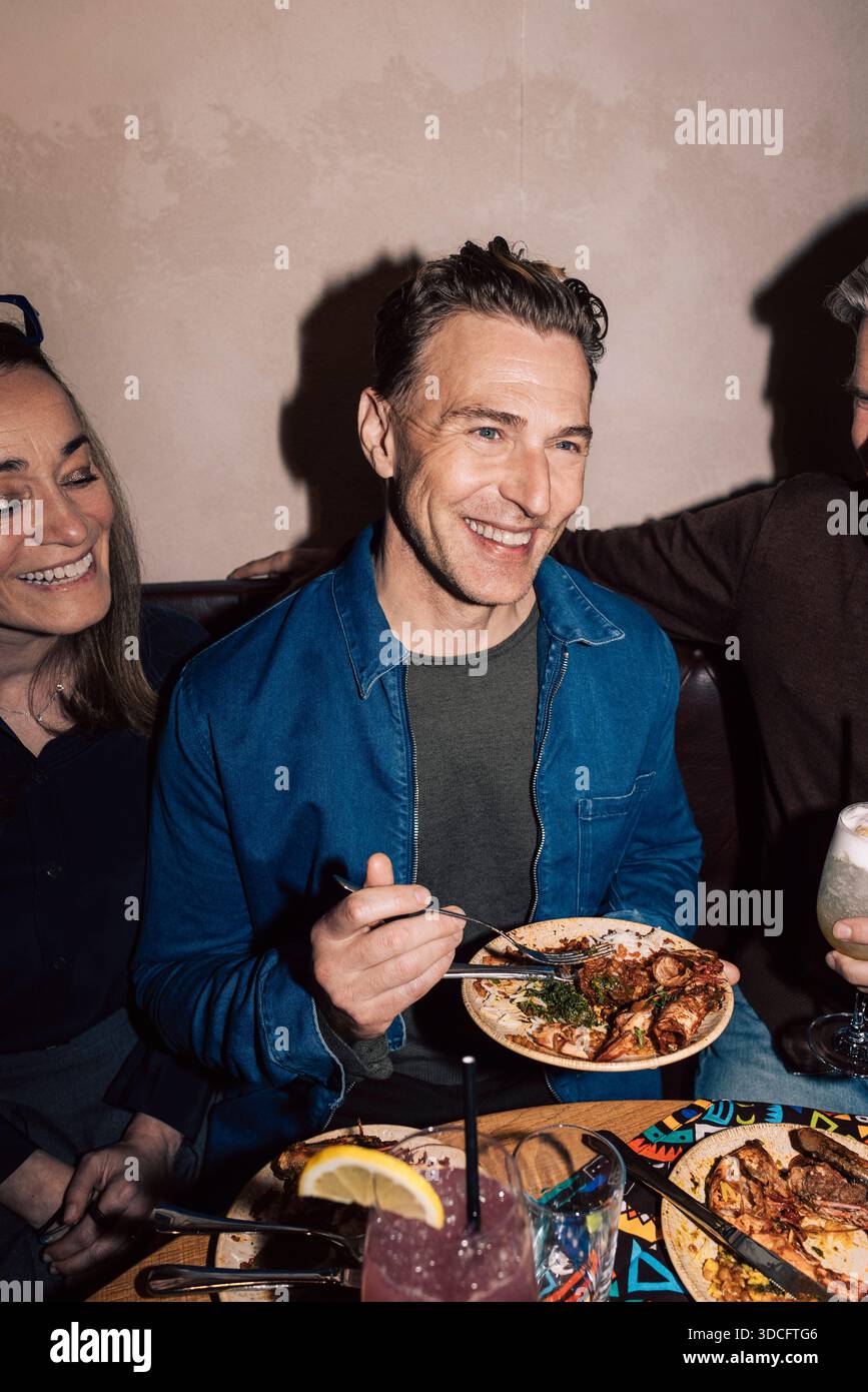 Uomo felice che mangia cibo mentre siede con gli amici a cena al ristorante Foto Stock