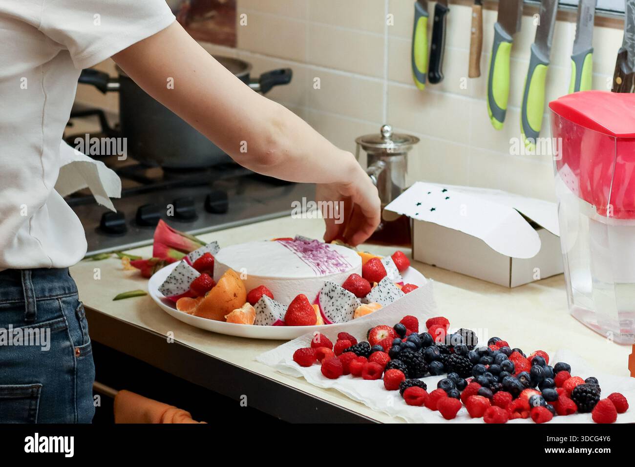 Vista laterale della persona in cucina che si avvicina per aggiungere frutti di bosco alla torta bianca con il condimento di frutta sul bancone Foto Stock