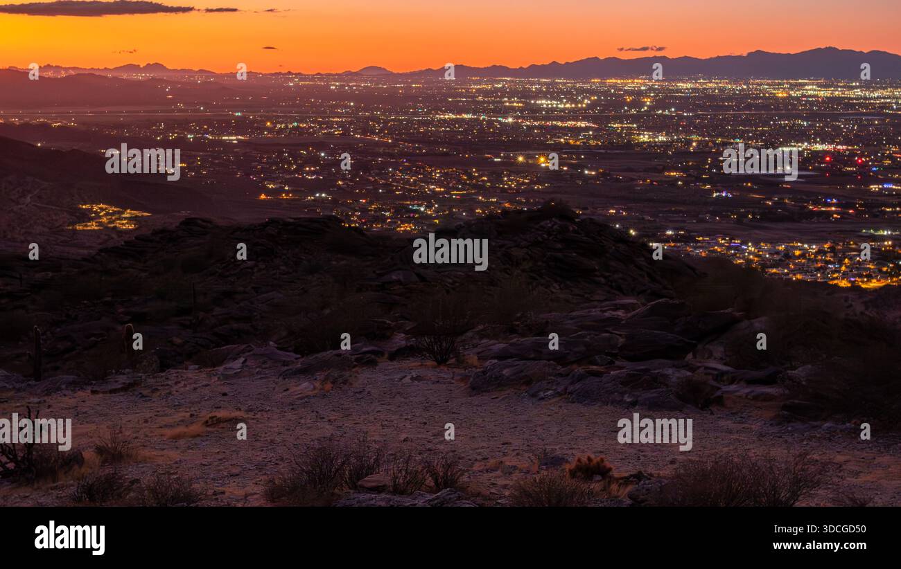 Vista di un paesaggio oscuro dove la sagoma scura del terreno roccioso incontra il vibrante bagliore delle luci della città sotto un fuoco cielo al tramonto, Phoenix, Arizona, Foto Stock