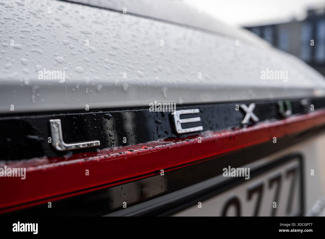 La vista ravvicinata ad angolo basso mostra le lettere cromate LEXUS sul rivestimento nero sopra una barra luminosa rossa, le gocce di pioggia fresche su vernice bianca, un frammento della targa e. Foto Stock