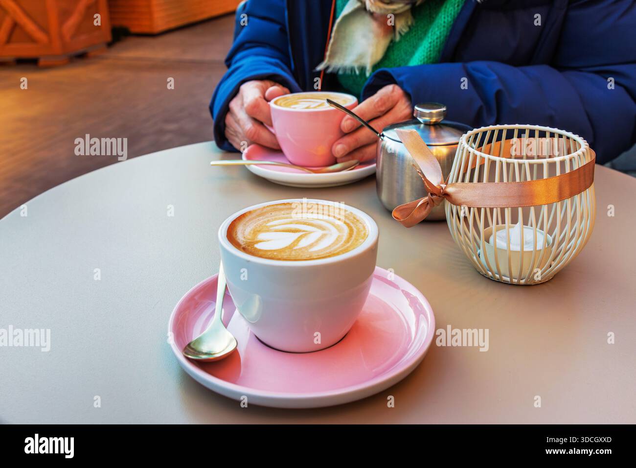 Due tazze di caffè con latte art si siedono su un tavolo da caffè con una lanterna a candela e una persona che tiene una tazza sullo sfondo. Foto Stock