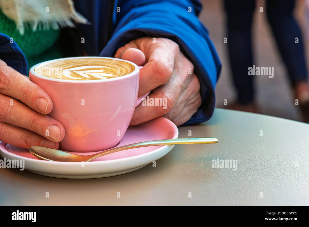 Una persona anziana tiene in mano una tazza bianca di cappuccino con latte art su un piattino rosa in un tavolino di strada in una giornata fredda. Foto Stock