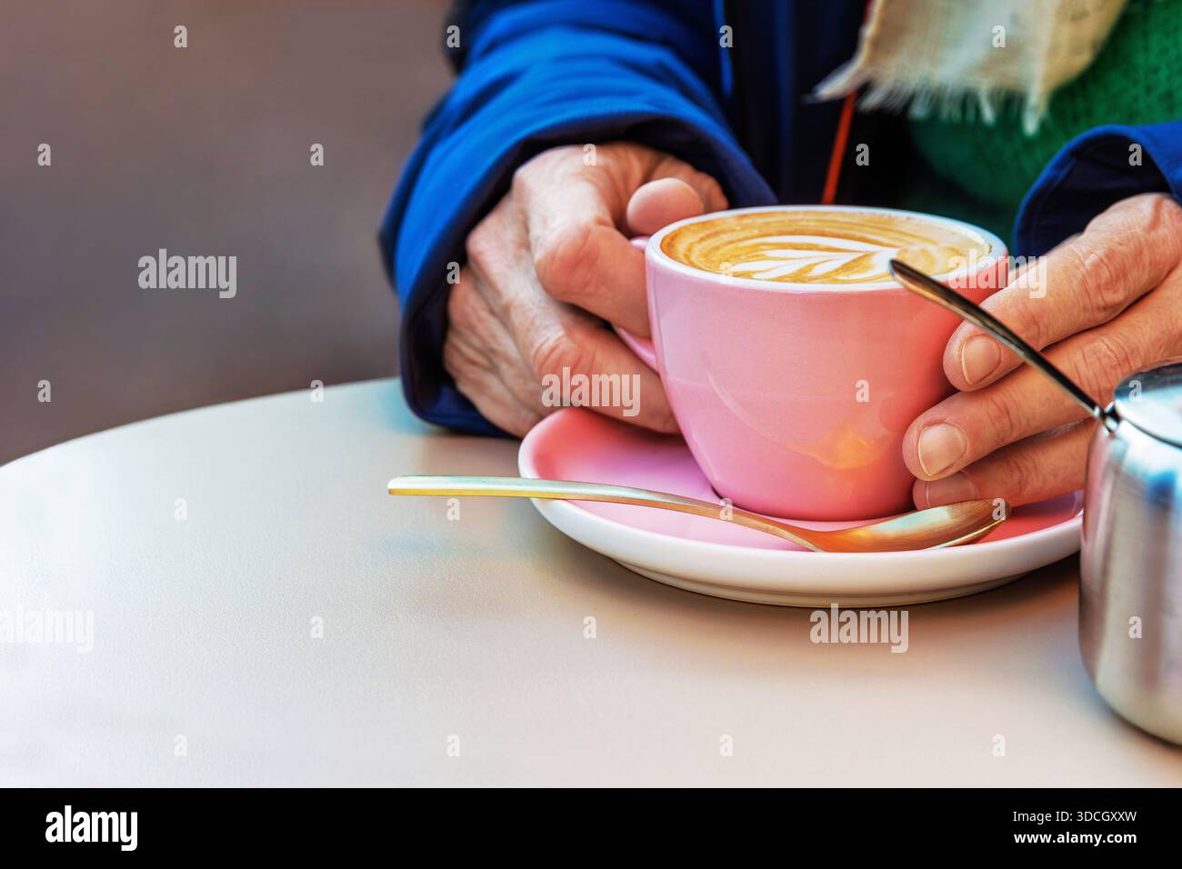 Una persona anziana tiene in mano una tazza bianca di cappuccino con latte art su un piattino rosa in un tavolino di strada in una giornata fredda. Foto Stock