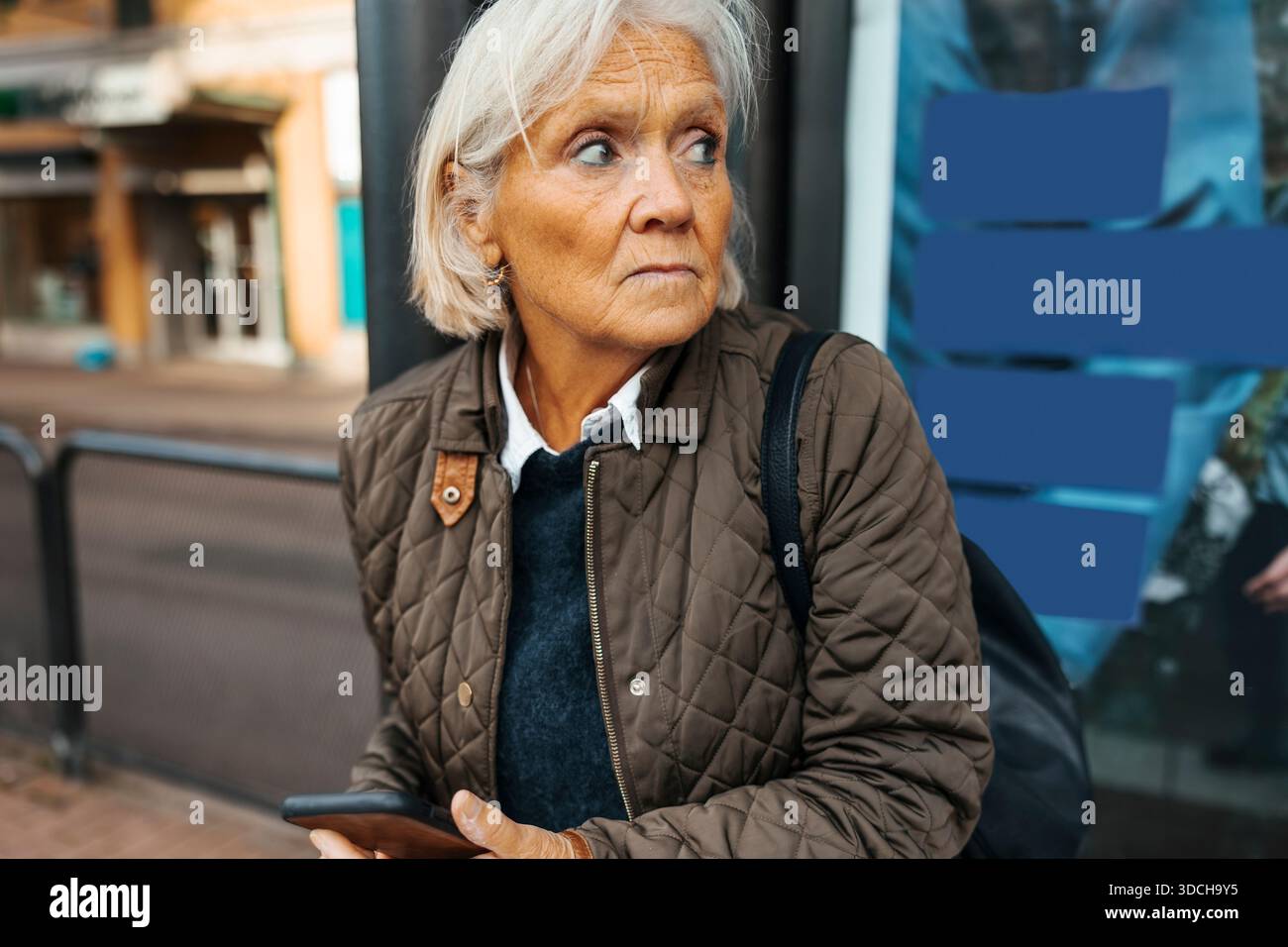Donna anziana dai capelli bianchi che guarda lontano mentre aspetta alla fermata dell'autobus Foto Stock