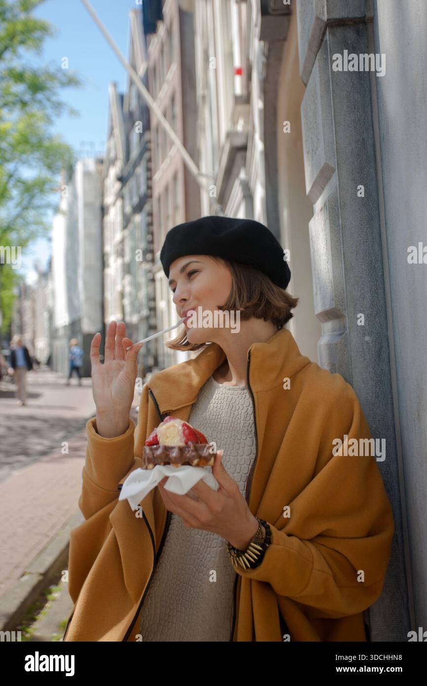 Bella ed elegante donna che indossa un berretto francese, appoggiata su un muro urbano mentre gusta un waffle con frutta fresca e cremosa. Foto Stock