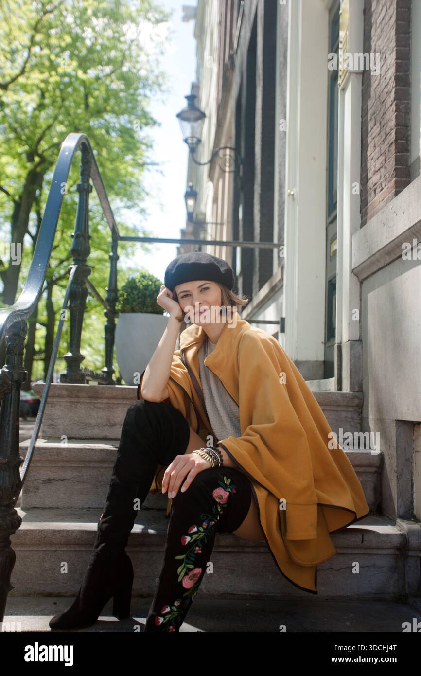Donna elegante che indossa stivali alti, cappotto elegante e berretto si siede sulle scale all'aperto in una giornata di sole. Stile di vita e viaggi con un'atmosfera affascinante. Foto Stock