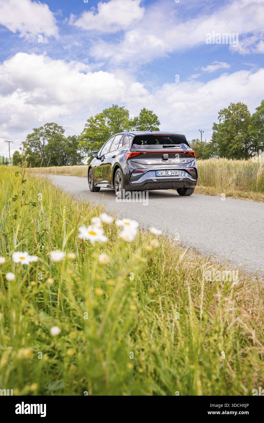 Auto nera che guida su una strada di campagna con prati in fiore e cielo nuvoloso, condivisione di e-car con cervi, auto elettrica Cupra, Germania Foto Stock