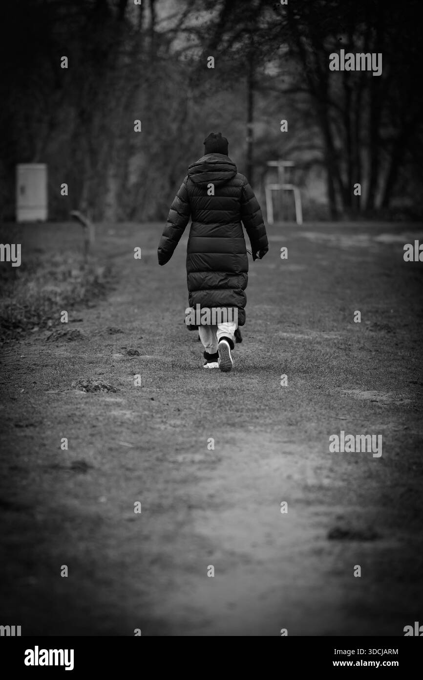 Una fotografia in bianco e nero di una persona solitaria che si allontana lungo un sentiero nella foresta durante una tranquilla giornata invernale. Il soggetto è vestito con una lunga imbottita Foto Stock