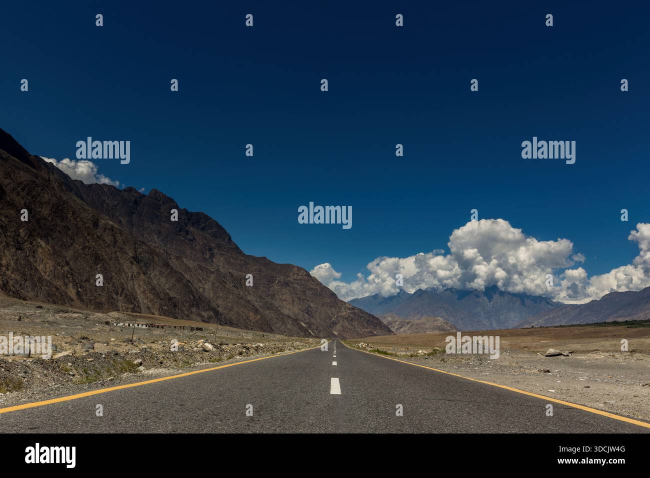 Vista di un nastro di asfalto senza fine che taglia attraverso il terreno roccioso e aspro sotto un cielo azzurro punteggiato da nuvole soffici, Jaglot, Gilgit Baltistan, P. Foto Stock