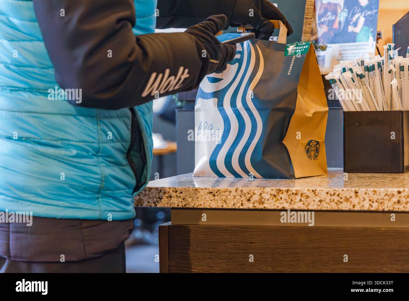 Un corriere per la consegna di alimenti Wolt in abbigliamento invernale raccoglie i sacchetti di carta Starbucks di marca da un banco di ritiro del bar Foto Stock