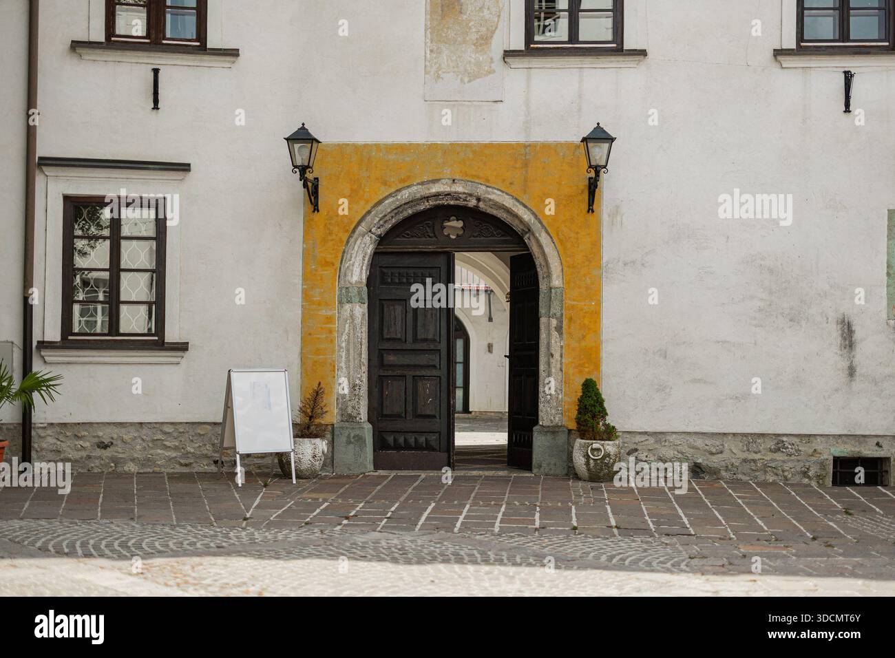 Un tunnel ad arco in pietra incornicia una vista di un edificio storico bianco con persiane marroni e un affresco religioso sbiadito. Una scala di legno è appesa Foto Stock