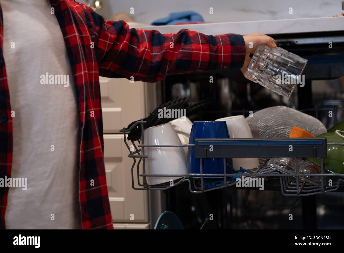 Primo piano di un bambino che prende un bicchiere pulito da una lavastoviglie aperta in una cucina di casa. L'immagine mostra la vita quotidiana della famiglia, le faccende domestiche e i bambini Foto Stock