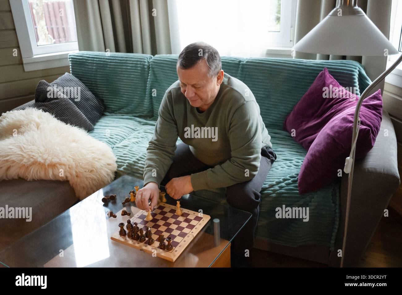 Uomo maturo seduto sul divano e giocando a scacchi da solo a casa, rilassandosi e trascorrendo del tempo in un accogliente salotto interno. Home attività ricreative Foto Stock