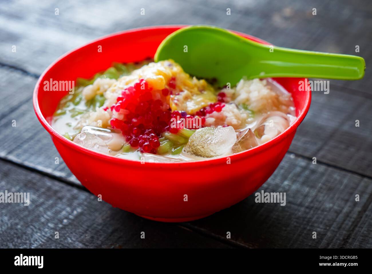 Cendol, un dessert malese servito in una ciotola rossa con ghiaccio tritato, gelatina verde, perle rosse, latte di cocco e riso glutinoso su un rustico tavolo di legno. Foto Stock