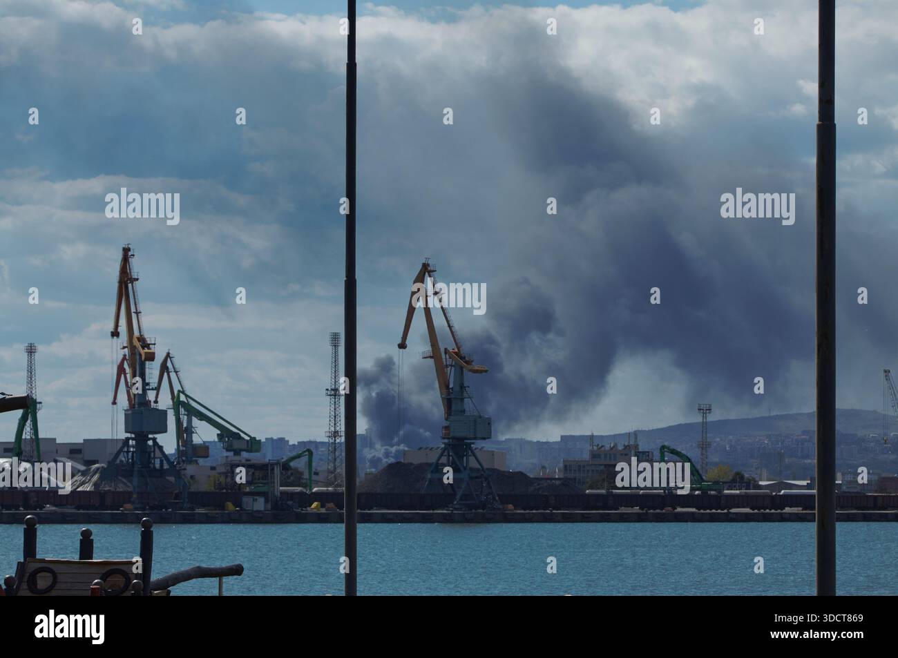 Il fumo sale da un incendio in un porto marittimo, con gru ed edifici industriali in primo piano, creando un'atmosfera di emergenza tesa e urgente Foto Stock